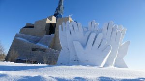A huge white sculpture of several white hands and another large grey sculpture behind it placed outside in the snow.